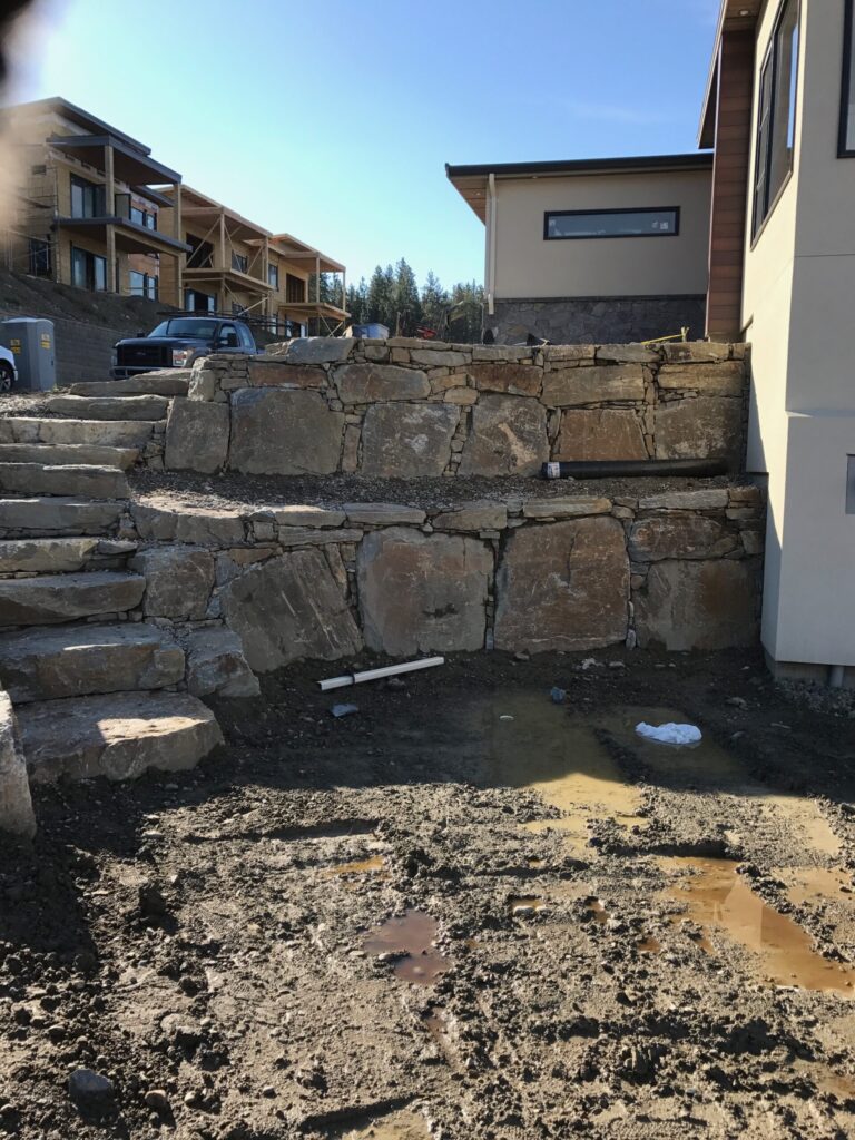 Construction site with a stacked stone retaining wall and unfinished multi-story buildings in the background; muddy, excavated ground in the foreground.